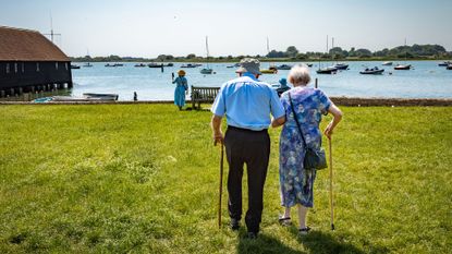 an elderly man and woman walk arm in arm and with canes towards a harbor full of sailboats
