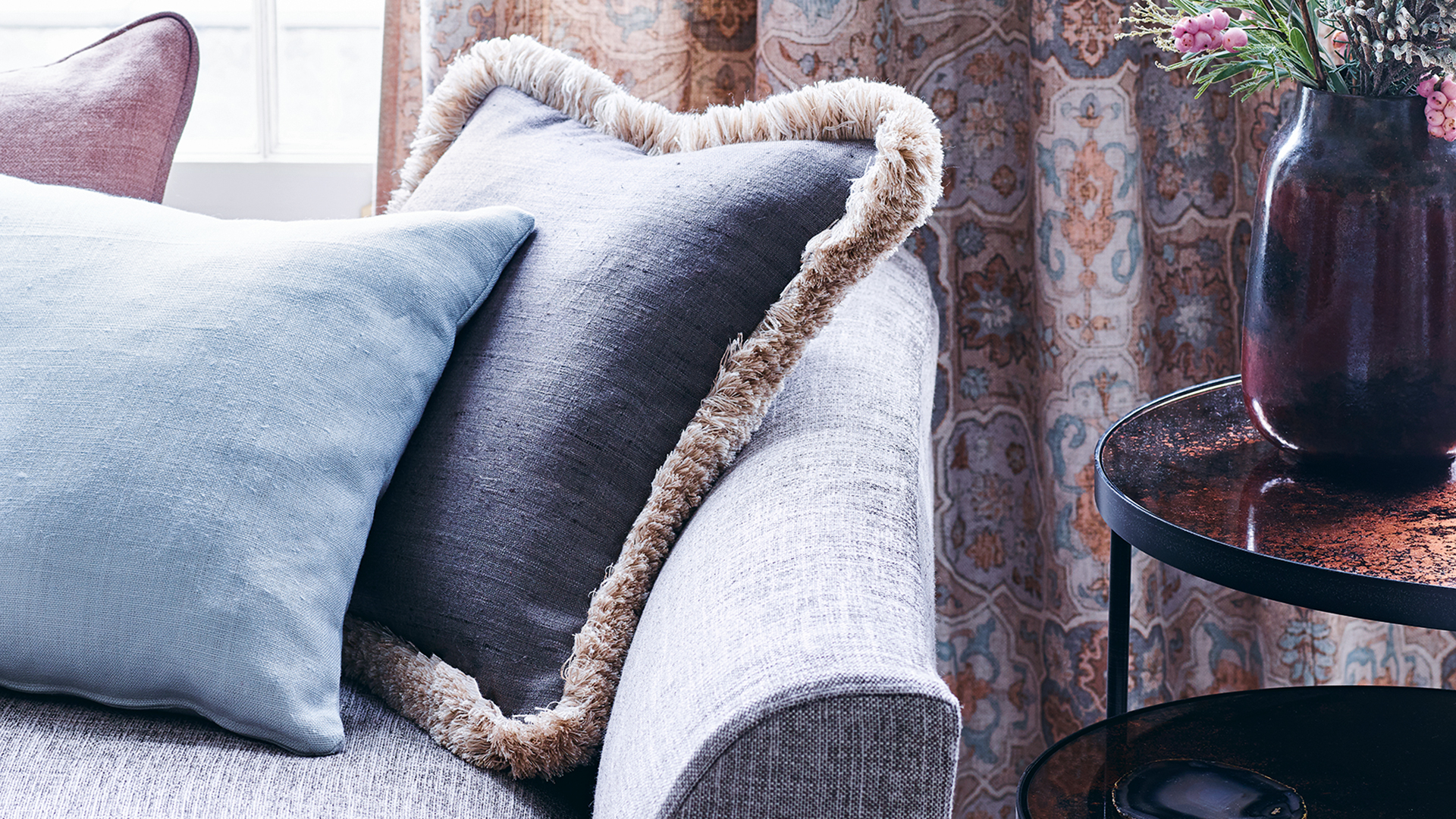 Close up image of a grey upholstered sofa with fringed cushions and pattern curtains in the background