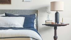 Neutral bedroom with white walls, and a blue and white patterned bed with matching bedding and cushions. Beside is a circular dark wood nightstand, dark blue lamp with white shade, and glass vase.