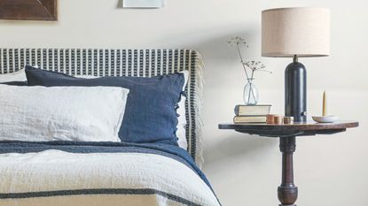 Neutral bedroom with white walls, and a blue and white patterned bed with matching bedding and cushions. Beside is a circular dark wood nightstand, dark blue lamp with white shade, and glass vase.