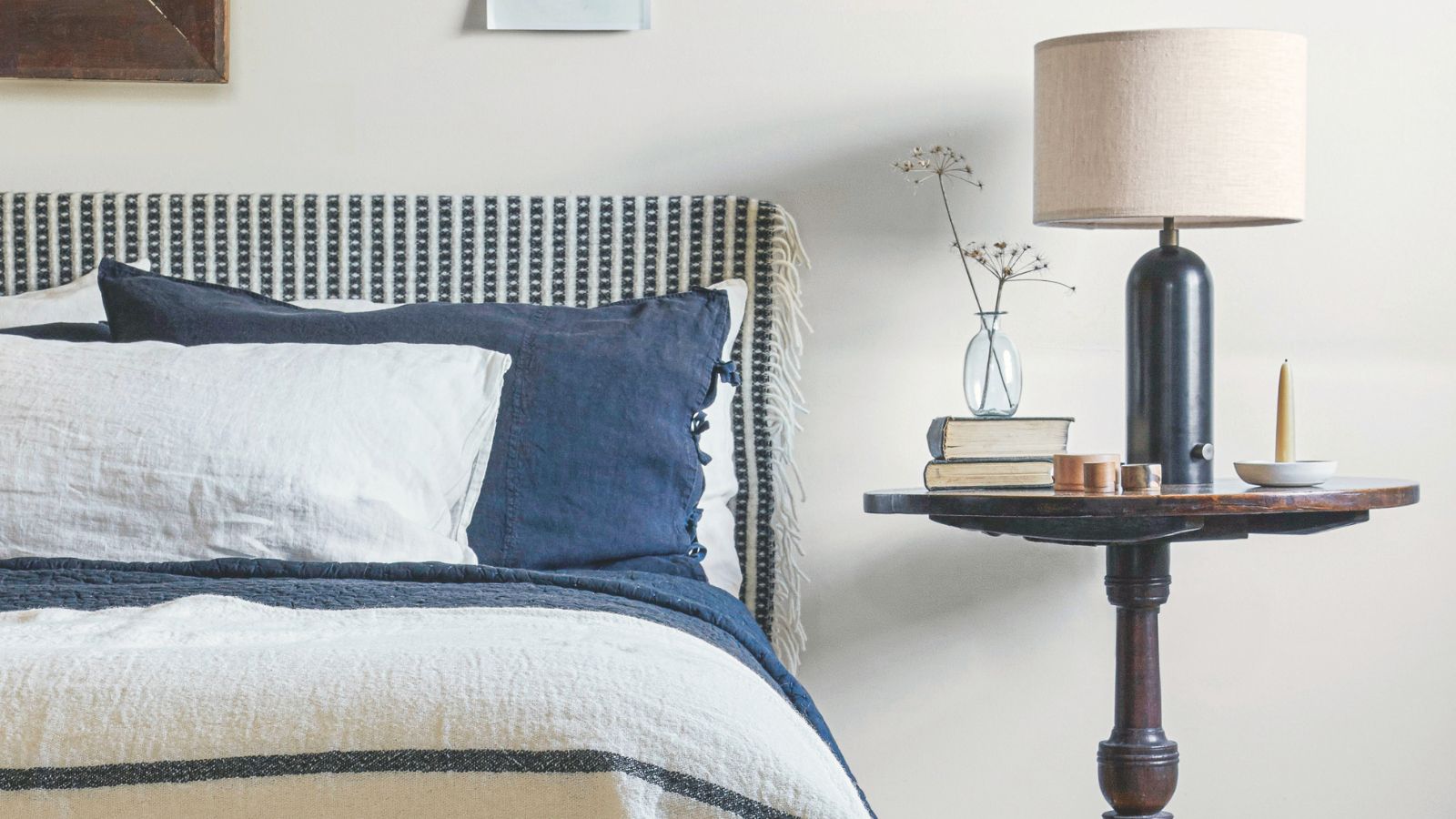 Neutral bedroom with white walls, and a blue and white patterned bed with matching bedding and cushions. Beside is a circular dark wood nightstand, dark blue lamp with white shade, and glass vase.