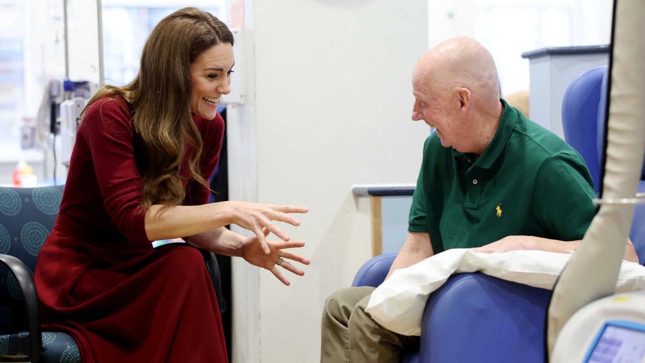 Kate Middleton wearing a red turtleneck and skirt sitting in a chair and talking and laughing with a man wearing a green polo hooked up to a cancer treatment machine