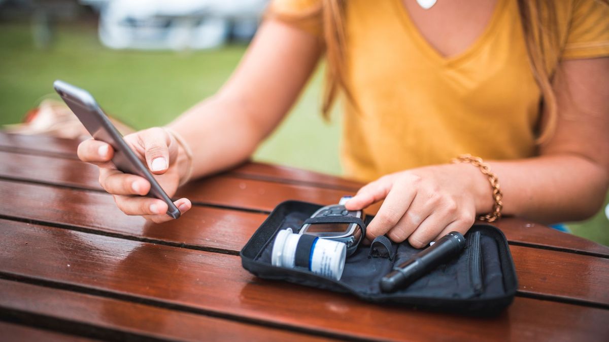 young woman in a yellow shirt uses her phone and a blood sugar monitor to check her blood sugar levels