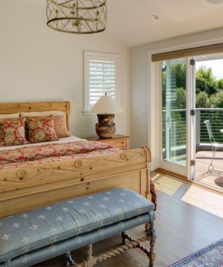 A farmhouse bedroom with a light wooden bed with a red floral throw pillows and a blanket on top of white sheets, a blue bed bench in front of it, and to the right an open door with a balcony overseeing green trees