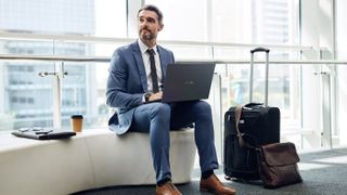 Man in suit sat on airport seating with laptop on knees and surrounded by luggage