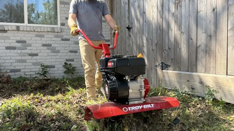 The Troy-Bilt 208cc 24 in tiller used on grass lawn by man with gloves, brown pants, and grey shirt