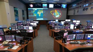 a flight control room. at the back are two large screens, one showing orbits around earth and the other showing a mission patch. in front of the screens are two rows of desks with computer screens on them