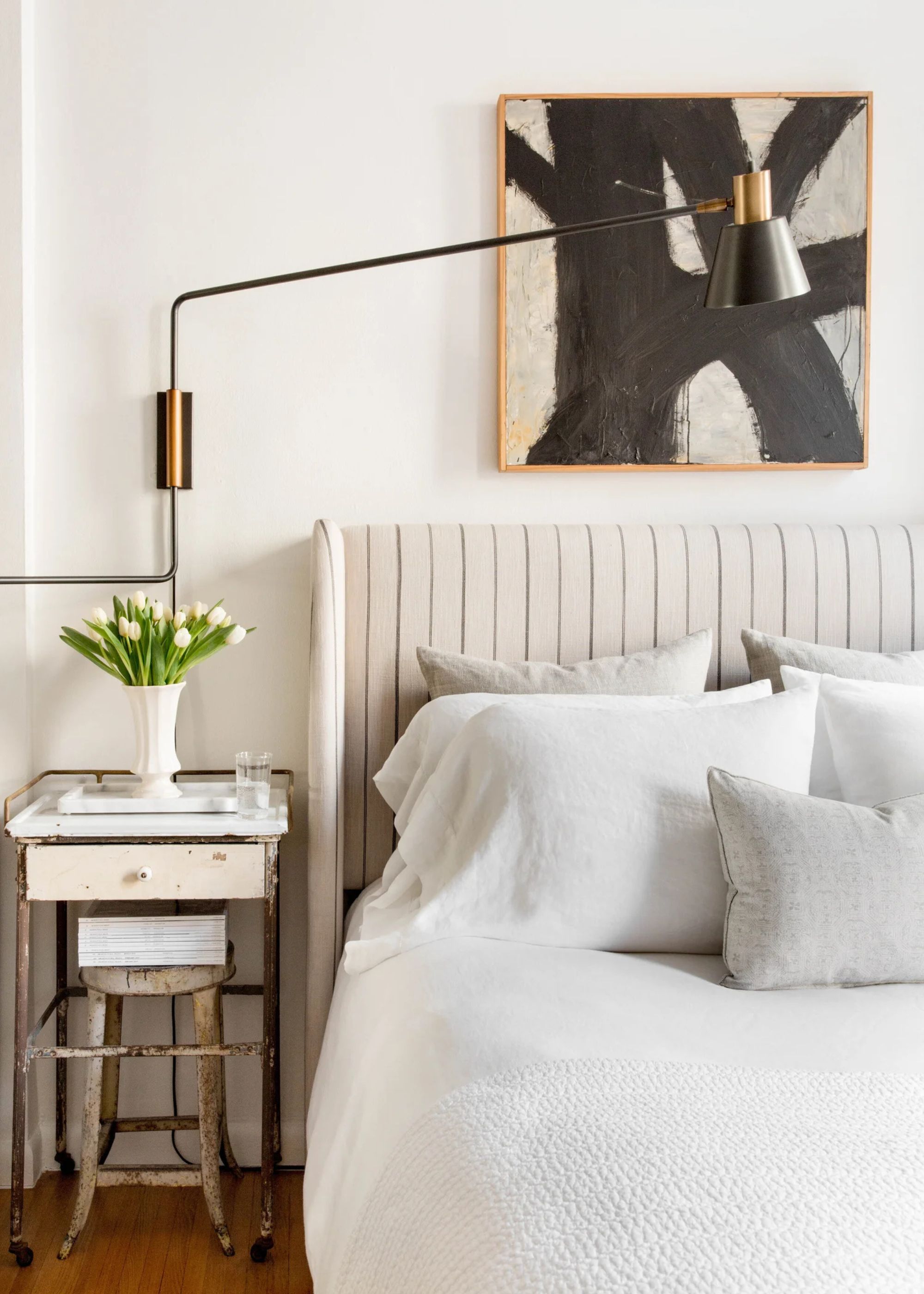 A small bedroom with white walls, a black abstract artwork, a bed with a neutral striped headboard and pale gray bedding, a vintage nightstand with a vase of flowers, and a black wall light.