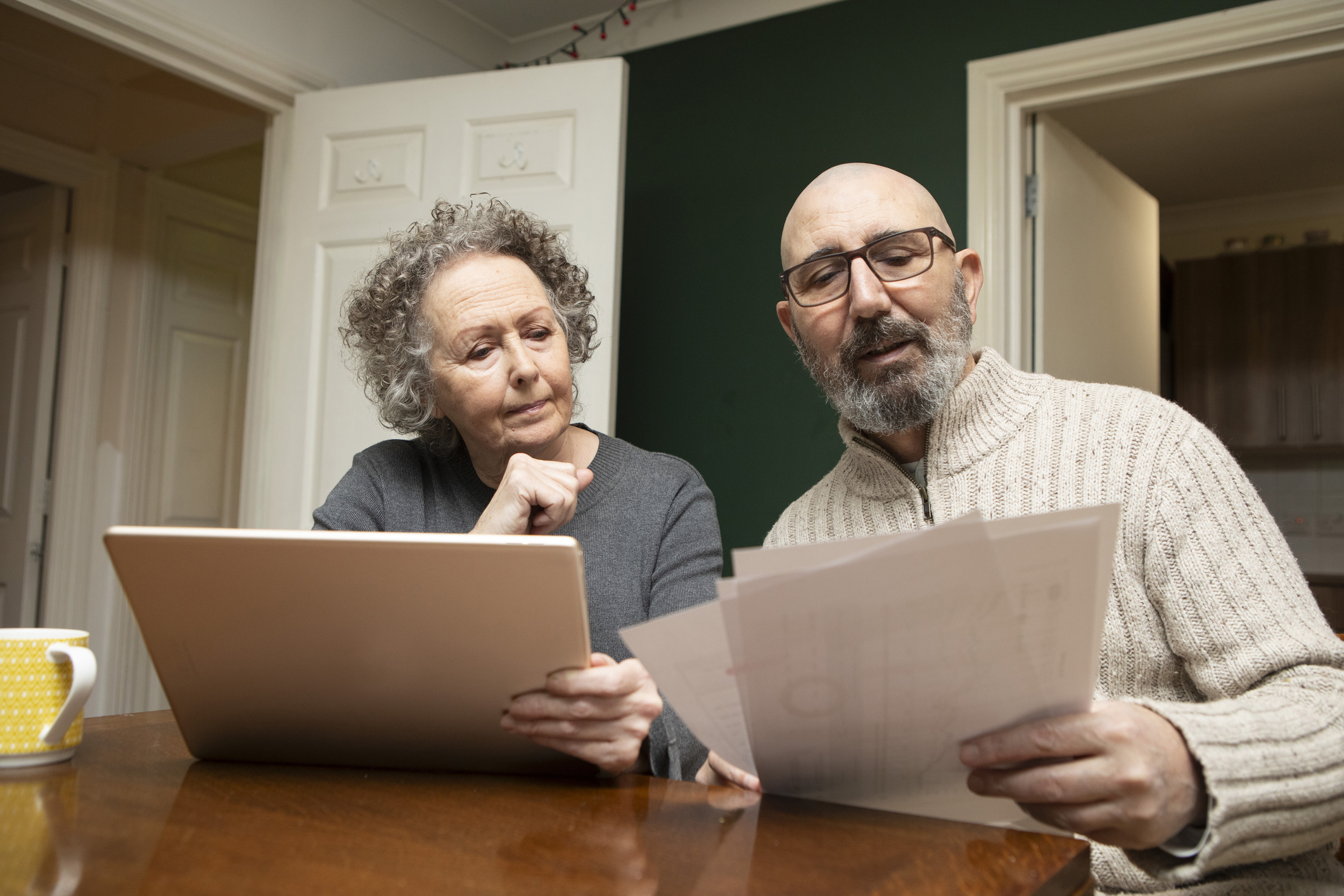 Senior couple at home reviewing financial information with a digital tablet and paperwork. They are both looking at the paperwork. They are casually dressed and sitting together at a wooden table.