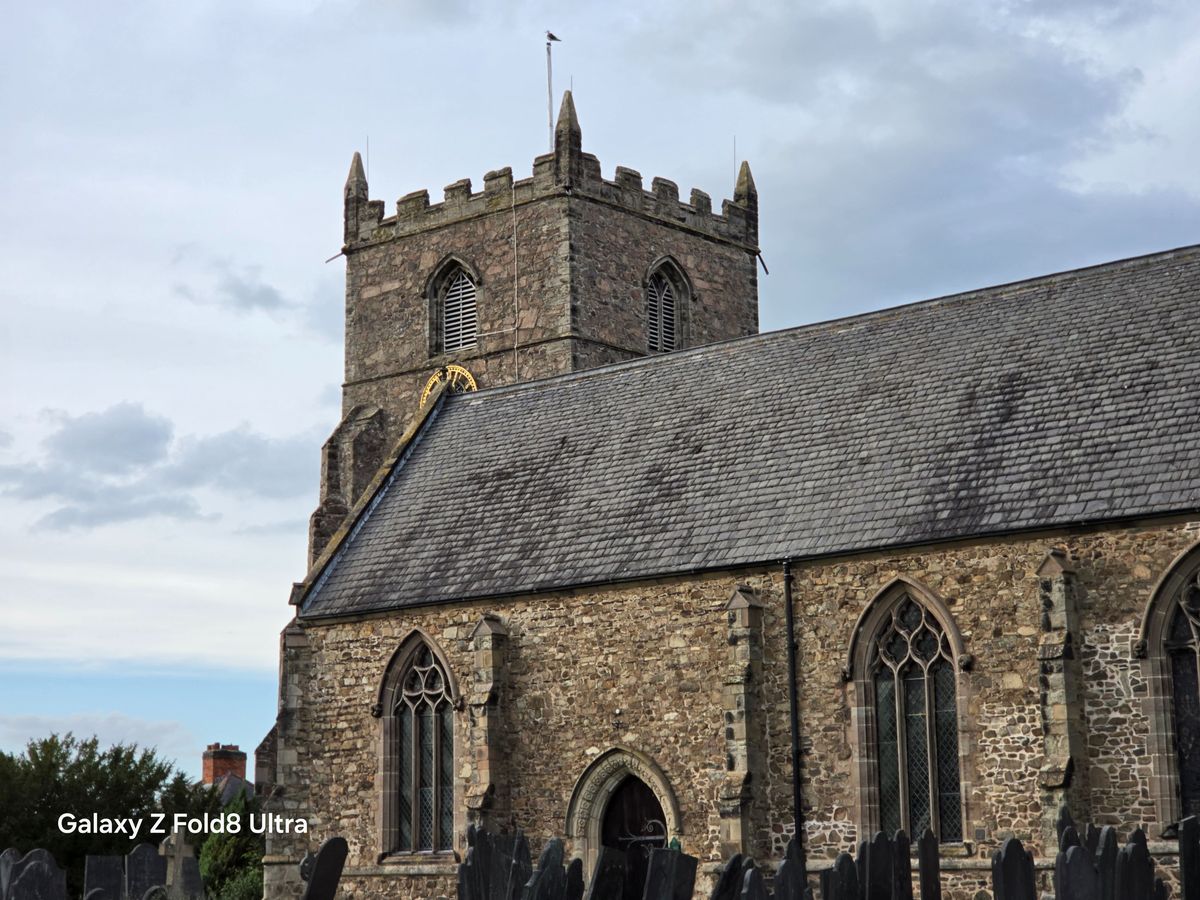 Telephoto view of a historic stone church with a square crenellated tower beneath a cloudy sky.