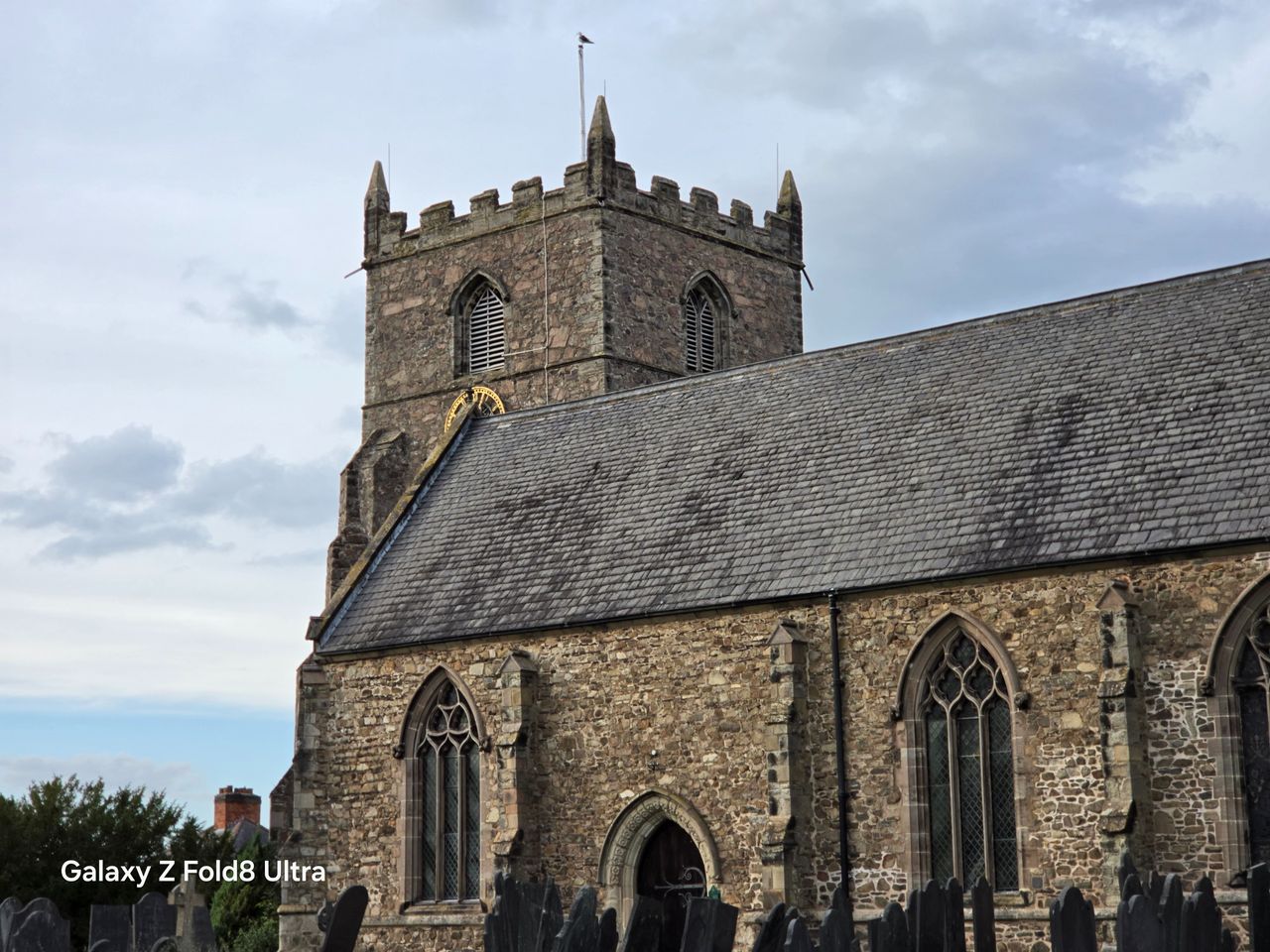 Telephoto view of a historic stone church with a square crenellated tower beneath a cloudy sky.