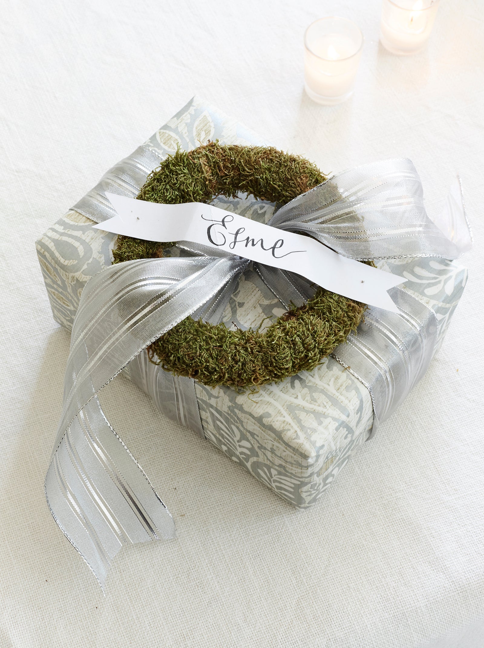 Christmas present wrapped in grey and white paper with silver ribbon, green moss wreath and name tag.
