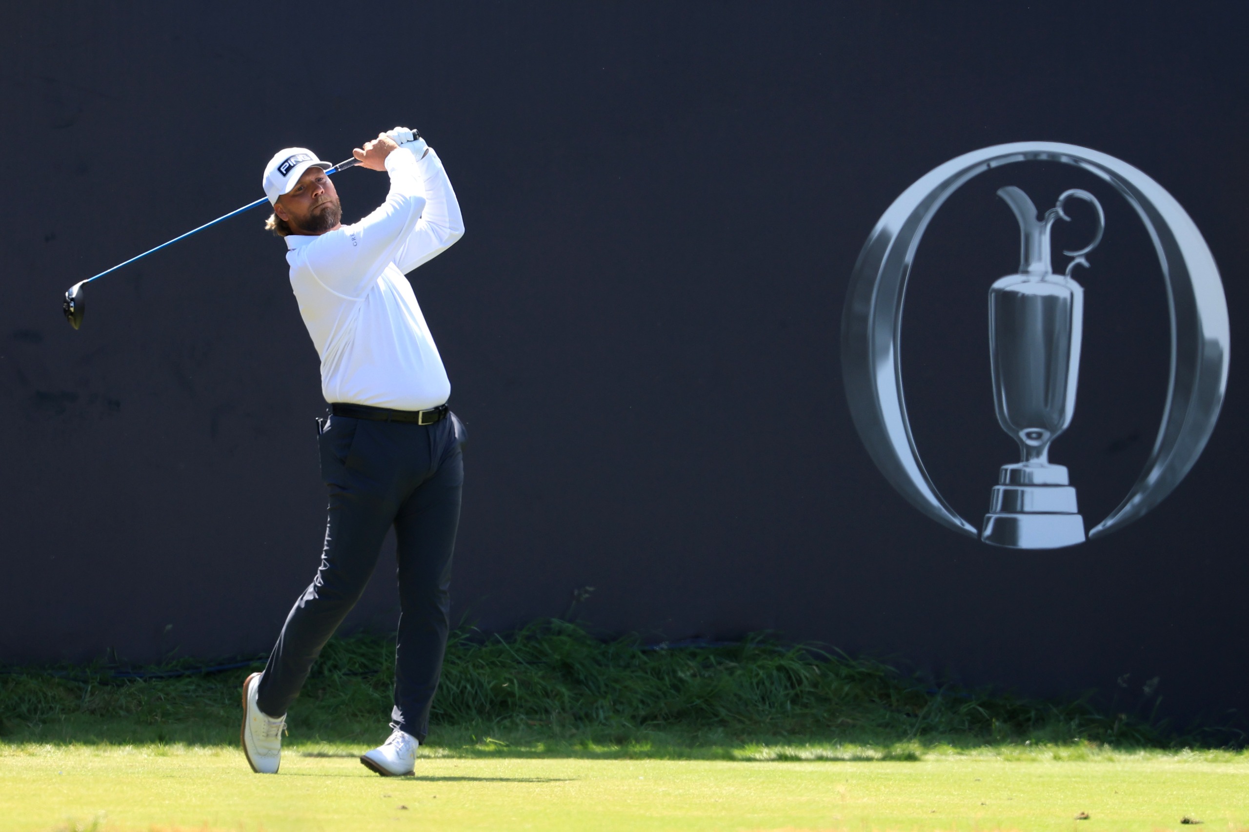 Dan Brown holds his finish on a drive at the 2026 Open in front of a blue board and claret jug logo