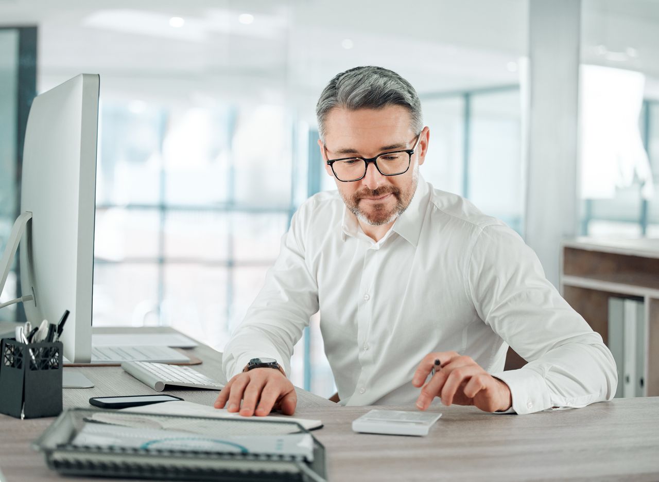 Man looks at calculator as he sits in front of computer screen at desk