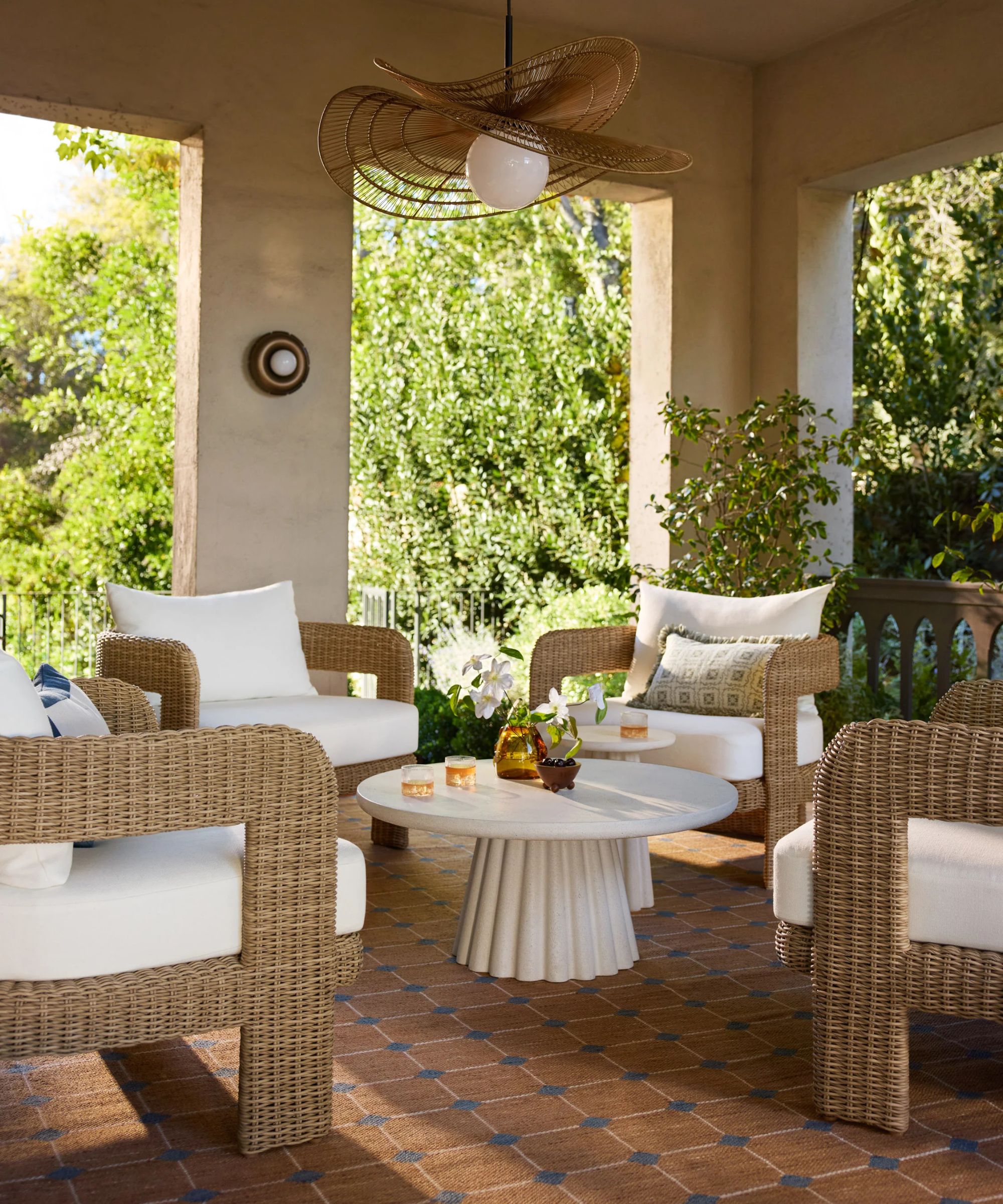 A bright, coastal-style patio featuring white lounge seating, a textured blue rug, and a large white coffee table. Several oversized woven rattan globe pendants hang from the ceiling, and a diamond-patterned trellis with climbing ivy decorates the back wall