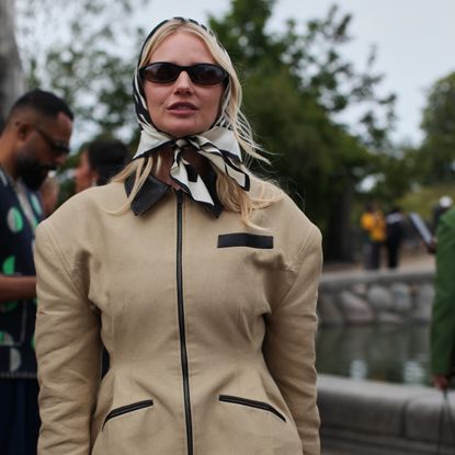 A Guest is seen wearing a beige barn jacket with a silk head scarf at the MKDT Studio show.