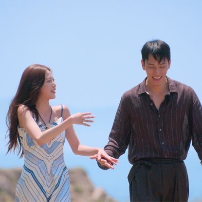A woman in a geometric-print dress (Lee Joo-young) and a man in a dark shirt and pants (Kim Jae-jin) hold hands and laugh while walking outdoors, with a rocky cliff and the sea behind them, in 'Single's Inferno' season 5.