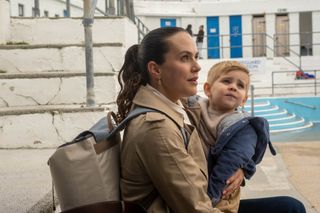 Lucy (Jessica Brown Findlay) is at a lido, carrying baby David in her arms and wearing a large backpack. She looks stressed and anxious.