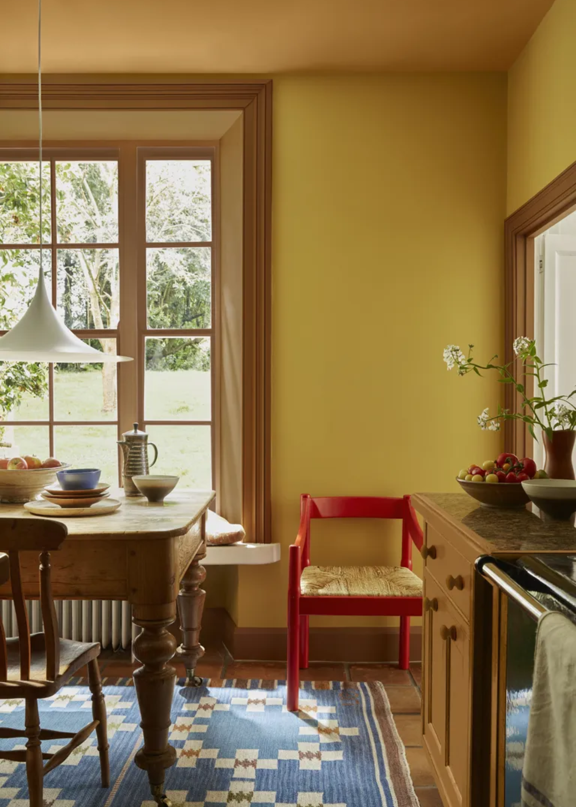 A dining room with a yellow ceiling, and mustard walls, a red chair, a patterned rug, and a wood dining set with a low-hanging white pendant lamp