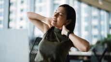 Woman sits in front of a computer in an office with her hands holding the back of her neck and her head tilted to the left