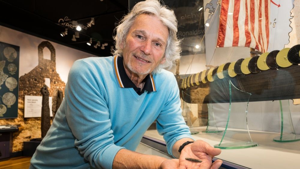 A man holds a silver ingot in the palm of his hand. He is in a museum with Viking artifacts, including a boat.