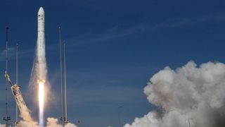 A white rocket blasts off against a blue sky. A plume of smoke rises on the right.