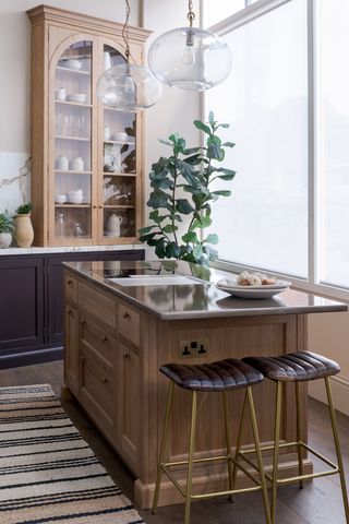 Beige kitchen with a light wooden kitchen island. There are two gold and brown leather bar stools, a stripped rug, and a large green plant in the corner. The island has a lava stone worktop, and there are two clear pendant lights above the counter