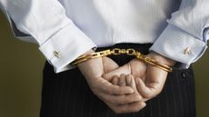 A business man with his hands cuffed behind his back with gold handcuffs.