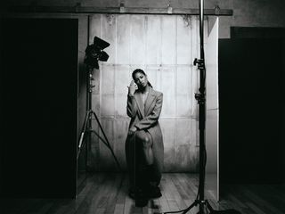 Black and white image of Lex Scott Davis in a studio setup sitting on a stool wearing a long coat.