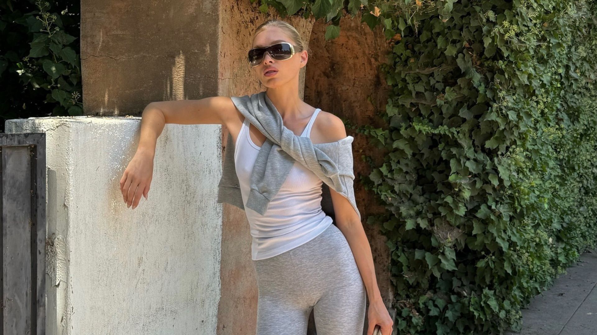 Model Elsa Hosk leans on a fence outside wearing grey capri pants with a white vest top, a Hermes bag and grey trainers styled with white socks.
