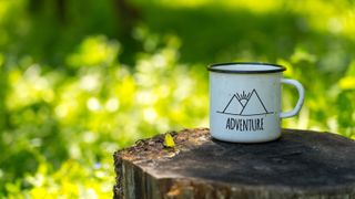 White enamel cup with mountains drawing and adventure text on aged stump in wild forest