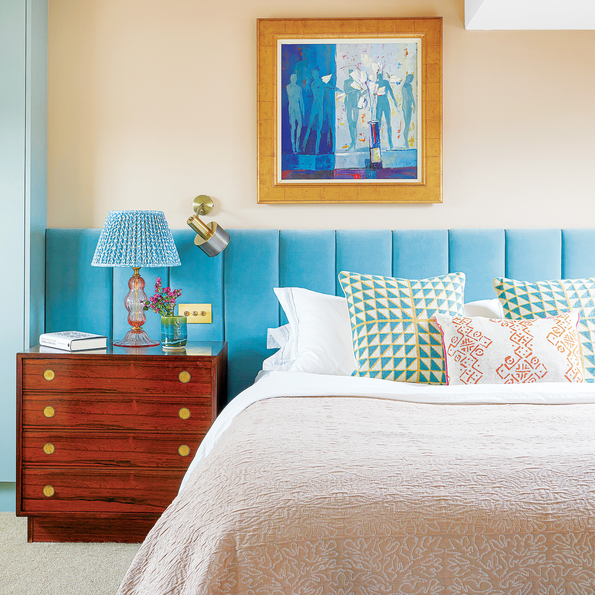 bedroom with cream walls and abstract artwork above cream and whtie bed and an oversized wide headboard in a sky blue velvet reaches the full width of the wall and behind a wooden bedside table 