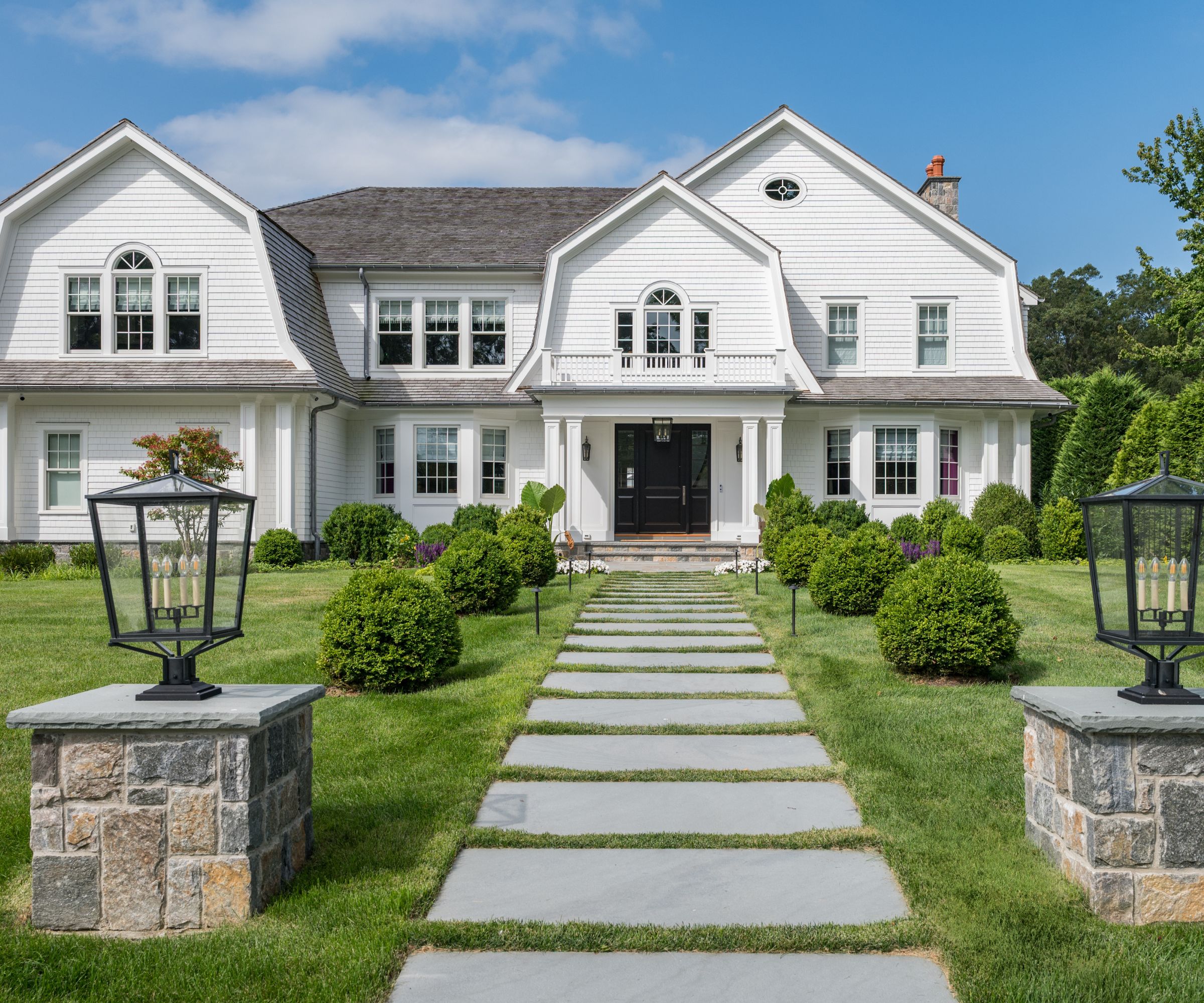 The exterior of a large traditional-style home painted white with a black front door and a large grass garden with shrubs and a paved path leading to the door.