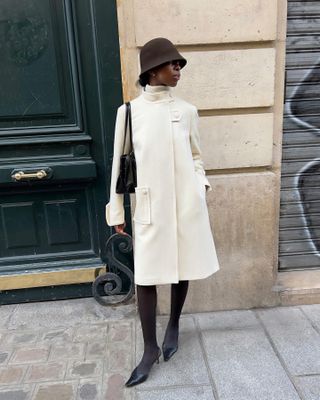 Sylvie Mus wearing a brown cloche hat, cream coat, tights, and black pumps.