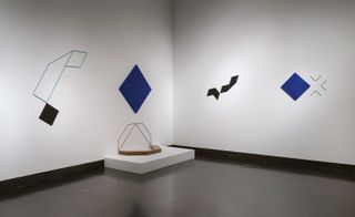 Installation view of Mehdi Moutashar’s black and blue geometric style works on the wall at the V&A - the space features white walls, black floors and a low white plinth displaying a piece of Moutashar’s art