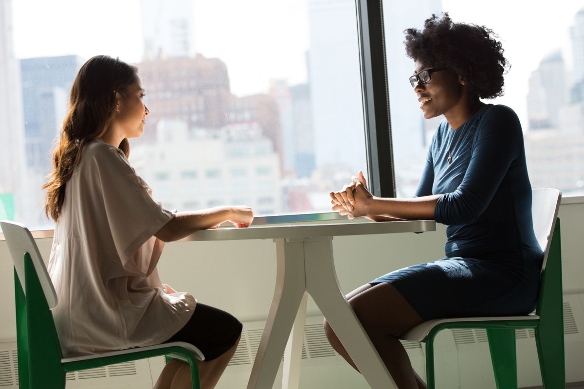 Two women sit talking at a table in front of a window overlooking a city