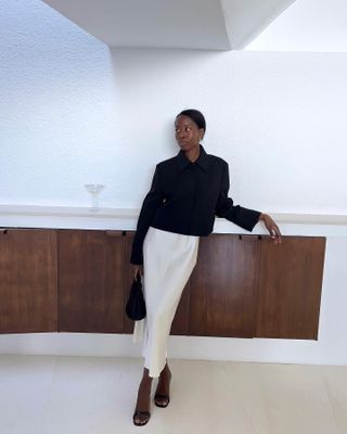 Sylvie Mus wears a black jacket, white slip skirt, black heels, and a black bag.