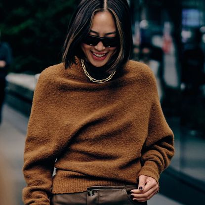 a woman at new york fashion week wearing a brown cashmere sweater, brown leather pants, and a gold chain necklace