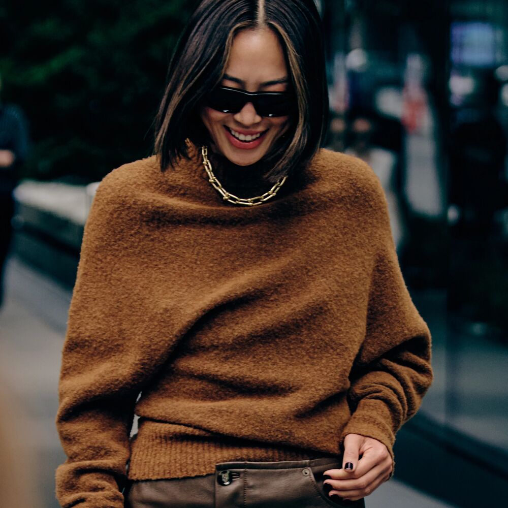 a woman at new york fashion week wearing a brown cashmere sweater, brown leather pants, and a gold chain necklace