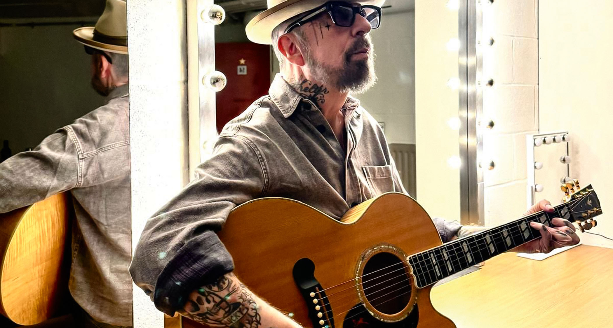 Dave Stewart in front of a dressing room mirror with a cutaway jumbo, wearing a wide-brimmed hat and shades.