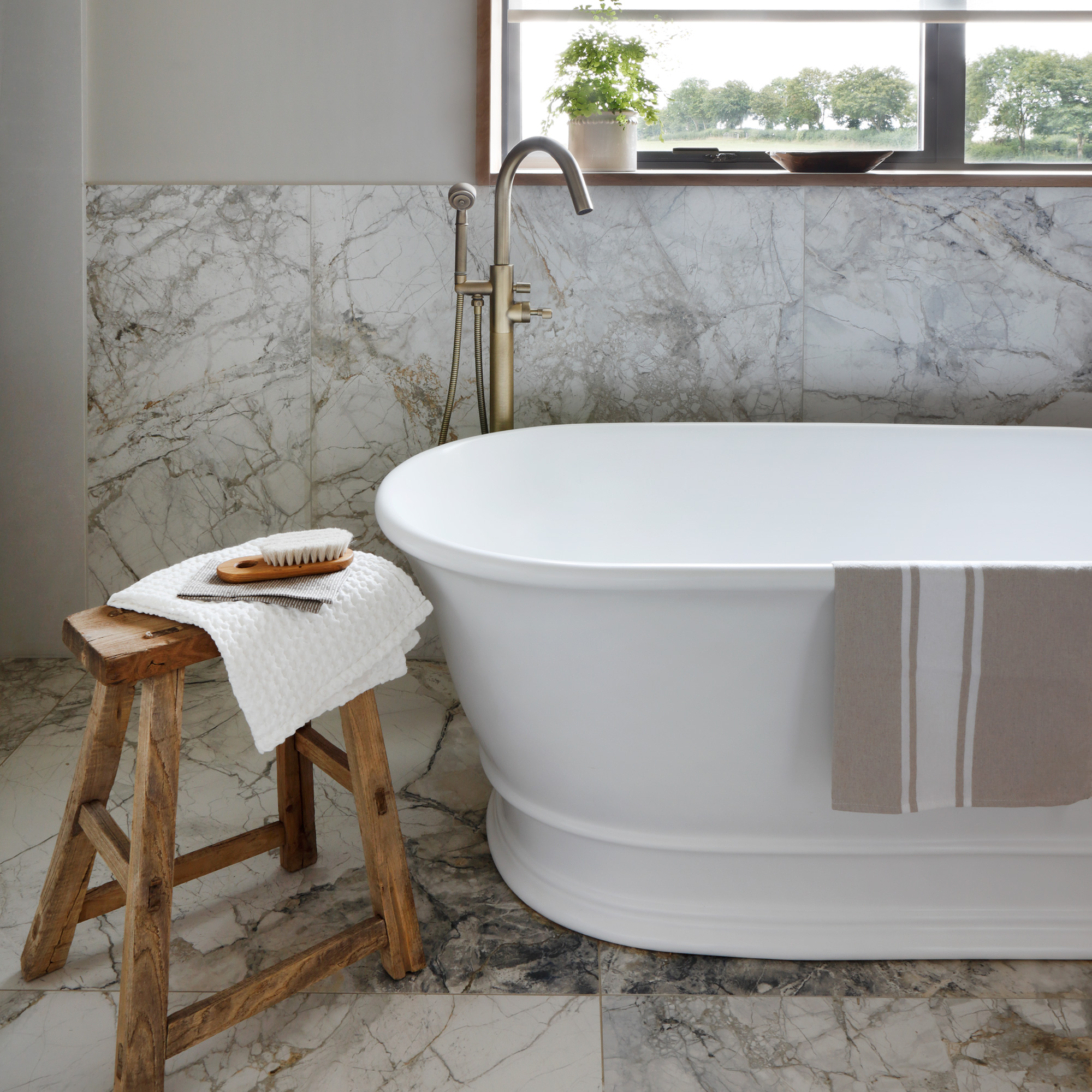 a neural bathroom with marble floor and wall tiles, a white bath tub and rustic wooden stool