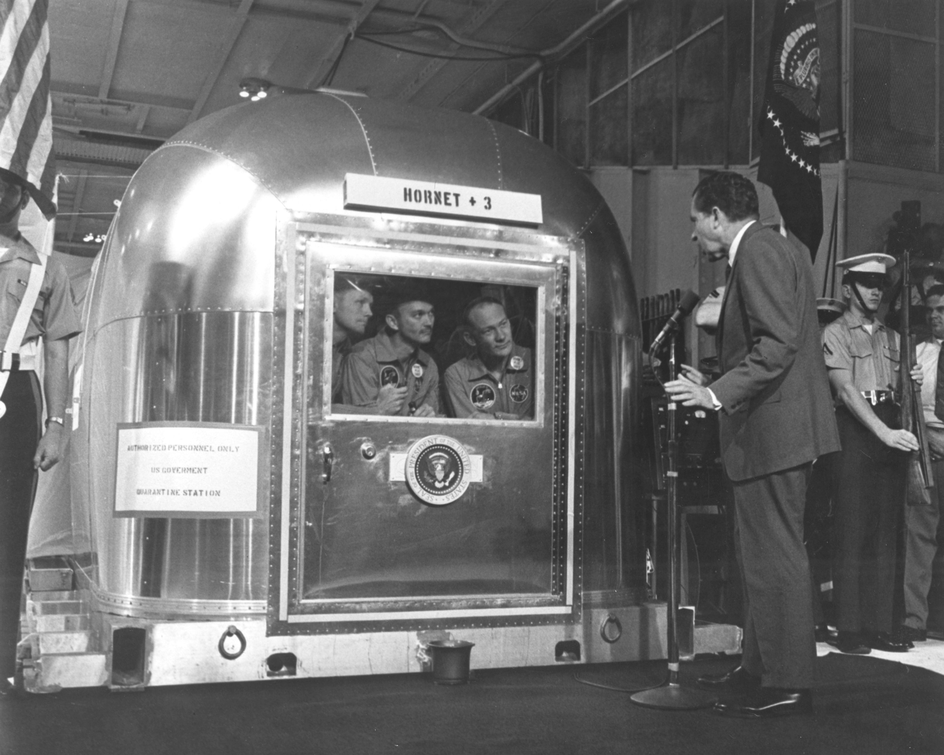 The three Apollo 11 astronauts peer out of a window in their metal quarantine trailer to greet President Richard Nixon.