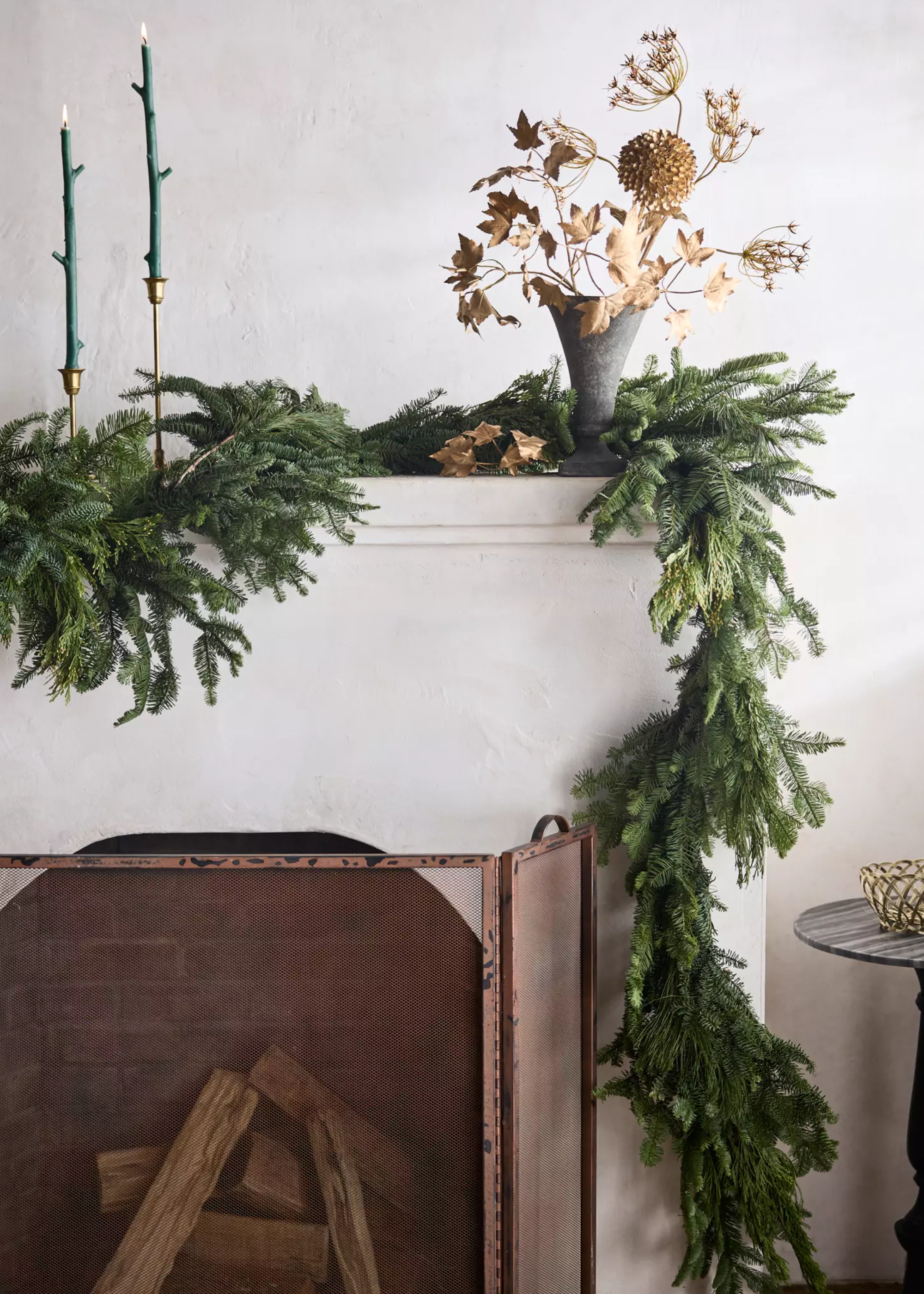 A Christmas garland on a mantle by green taper candles and a vase of gold flowers