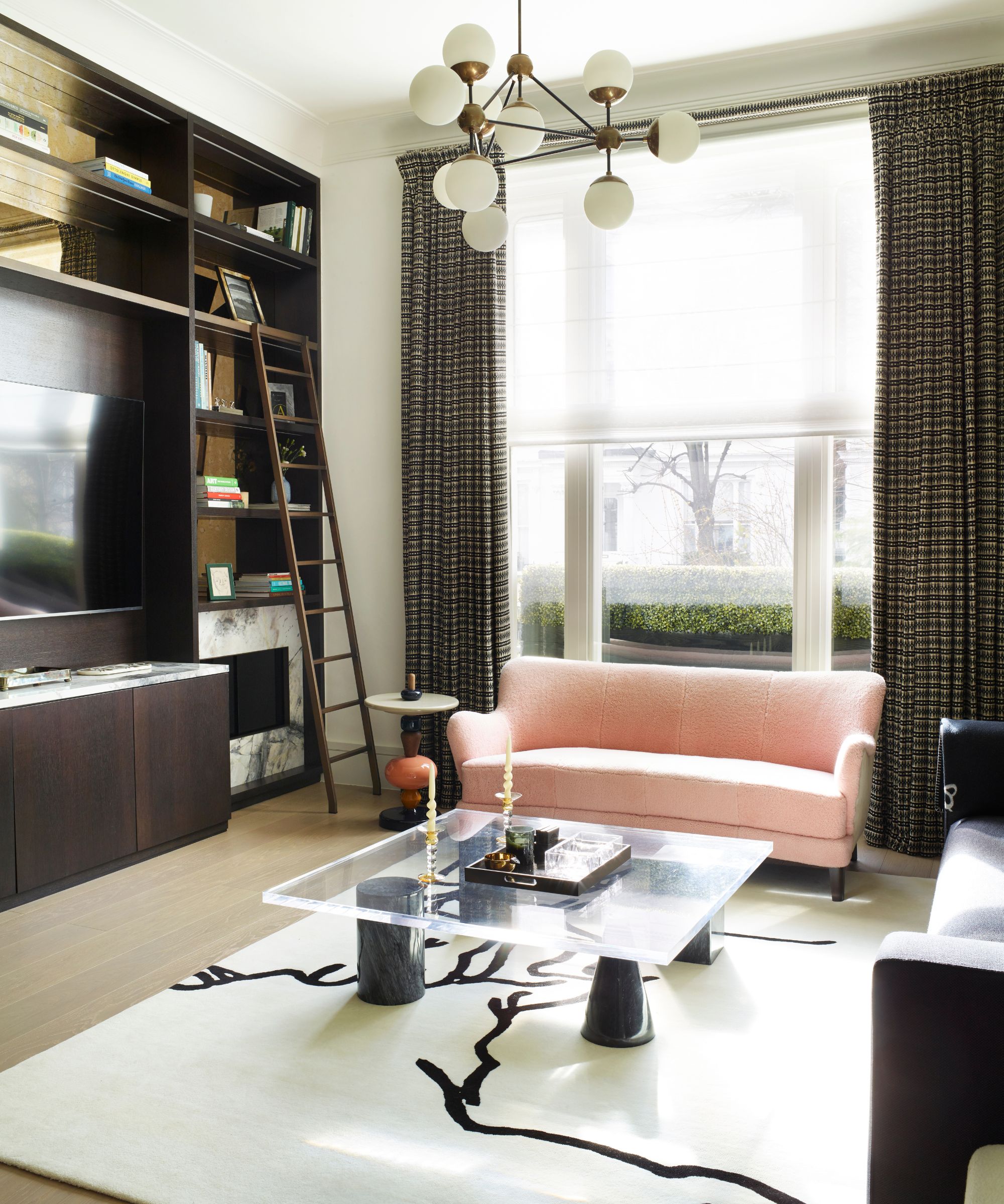 A bright living room with a large white statement rug, statement gold light fixture, glass coffee table, pink couch, wooden bookshelves with a ladder and TV, and large windows behind with striped curtains.