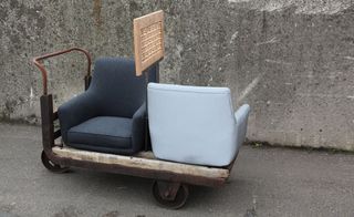 Concrete floor and walls, wooden frame trolley on metal wheels, with two back to front sitting chairs in shades of blue and grey on top, metal handle, wooden sign on a metal stand in-between the chairs