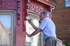 An older man in an historic town paints his house, with careful attention to detail, while standing on a ladder.
