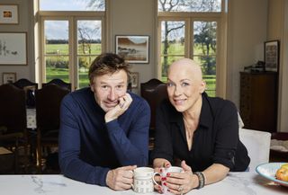 Sarah Beeny Vs Cancer: Sarah Beeny and her husband Graham Swift sit at their dining table, smiling, each of them resting a mug on the table. Sarah is bald in the picture, and we can see the views of their country estate through the windows behind them