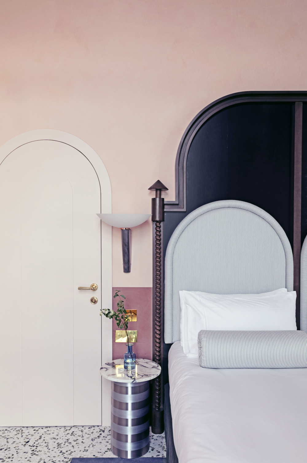 A rose-tinted hotel room features whimsically shaped banquette, coffee tables, and sconces, along with two-shade-painted walls and terrazzo floors.