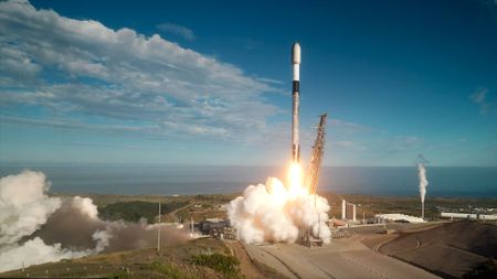 A white, black and gray-ish (soot-covered) rocket lifts off into a mostly-blue sky from a sea-side launch pad.