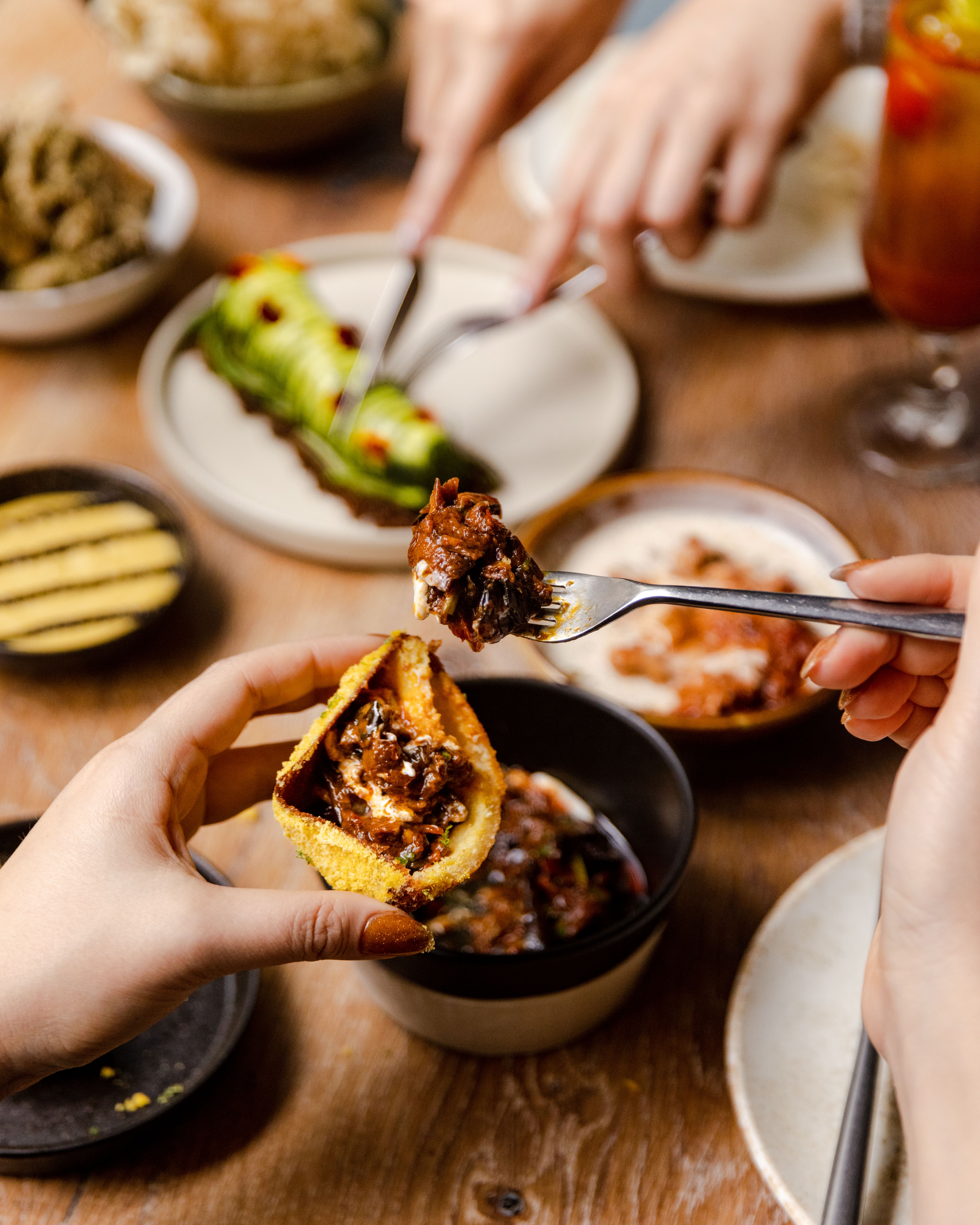 A seen-from-the-above dining table appears covered in multiple sharing platters filled with colorful dishes that are being handled by manicured hands of women.